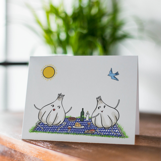 Card with illustration of two garlic bulb at a picnic on a wooden surface with a plant in the background