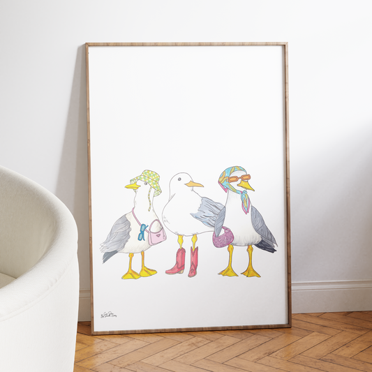A framed art print leaning against a white wall on a wooden parquet floor. The print features a whimsical illustration of three seagulls standing together, each with a different colorful hat and boots.