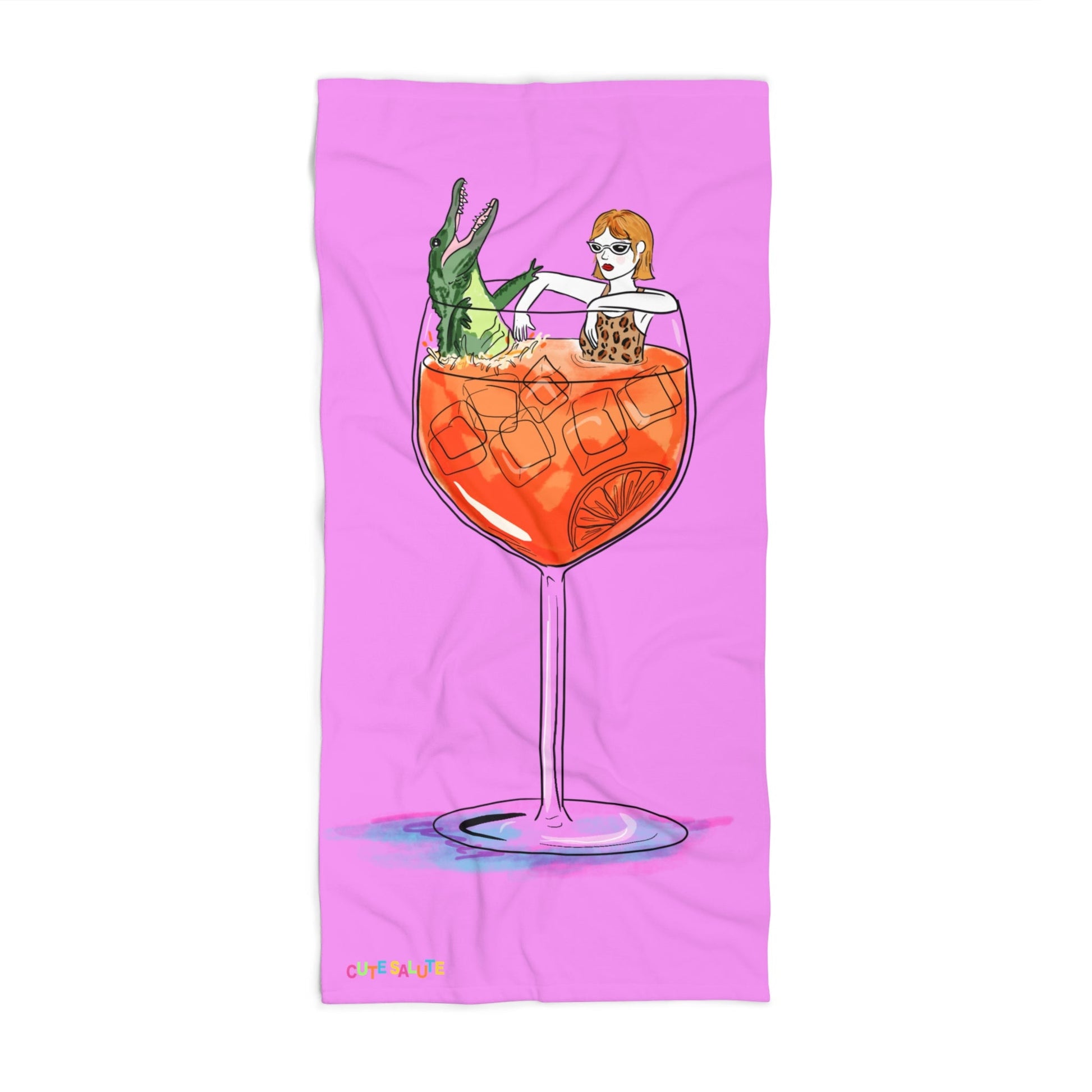 A pink rectangular towel featuring a whimsical illustration of a woman with orange hair sitting inside a large orange cocktail glass with an alligator, shown on a white background.