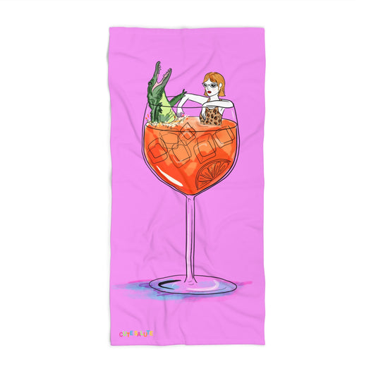 A pink rectangular towel featuring a whimsical illustration of a woman with orange hair sitting inside a large orange cocktail glass with an alligator, shown on a white background.