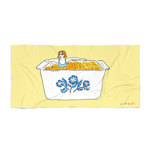 A pale yellow rectangular towel or mat with a printed illustration of a woman sitting in a corningware blue cornflower dish dish filled with yellow-macaroni and cheese. The towel is shown flat against a white background.