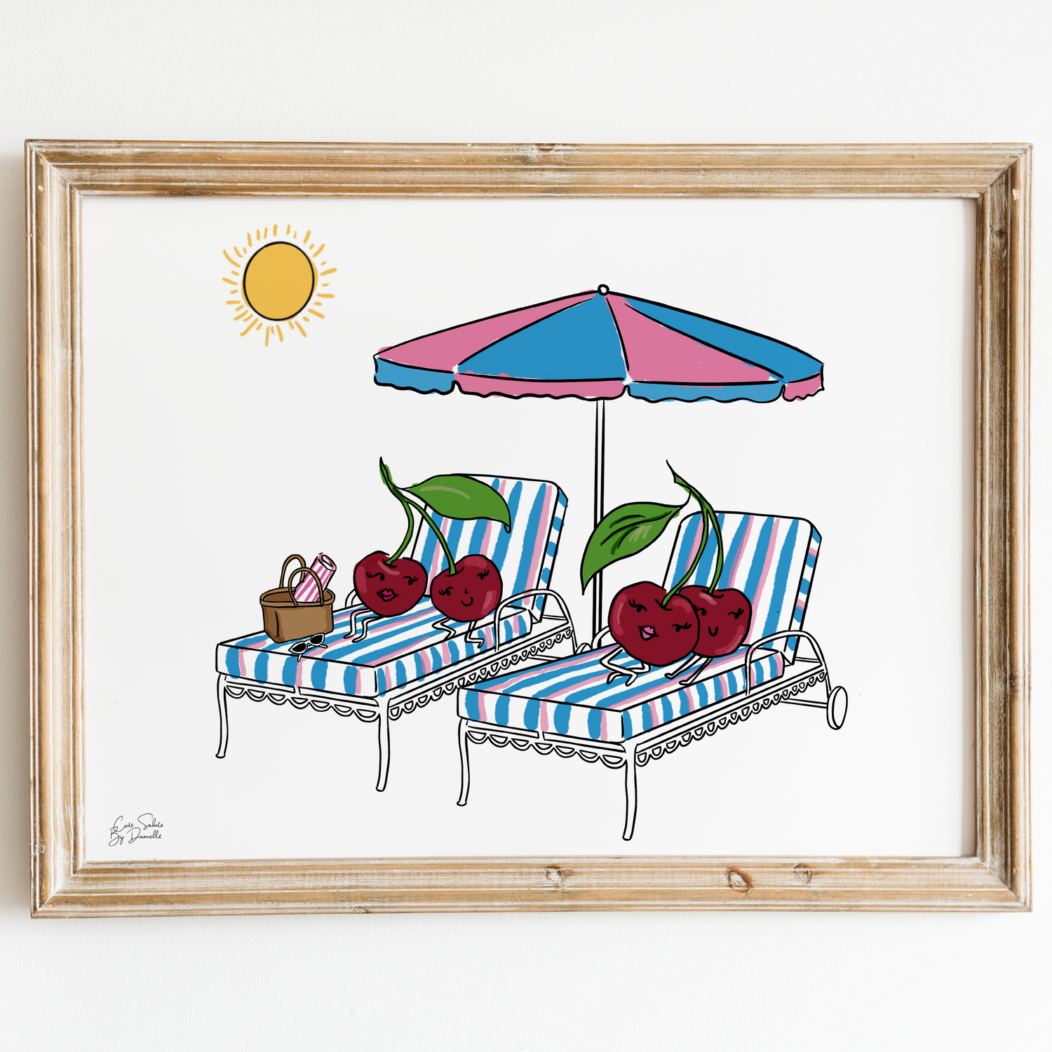 A framed art print featuring a whimsical illustration of two lounge chairs under a pink and blue striped umbrella, decorated with red cherries, on a white background with a light wood frame.