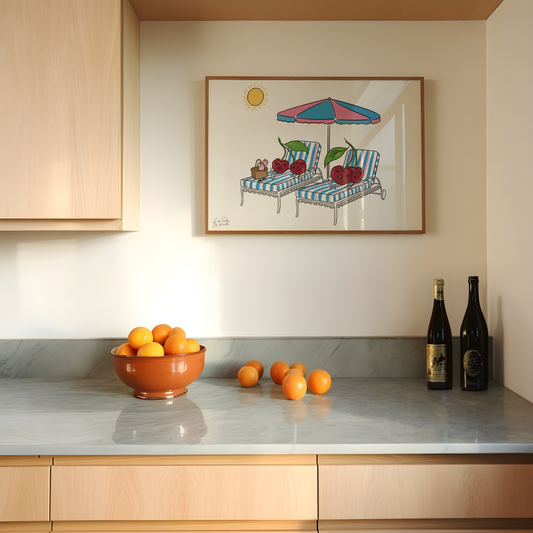 A framed art print depicting a sunny beach scene with striped lounge chairs and cherries, hanging on a beige wall above a kitchen counter with oranges and wine bottles.