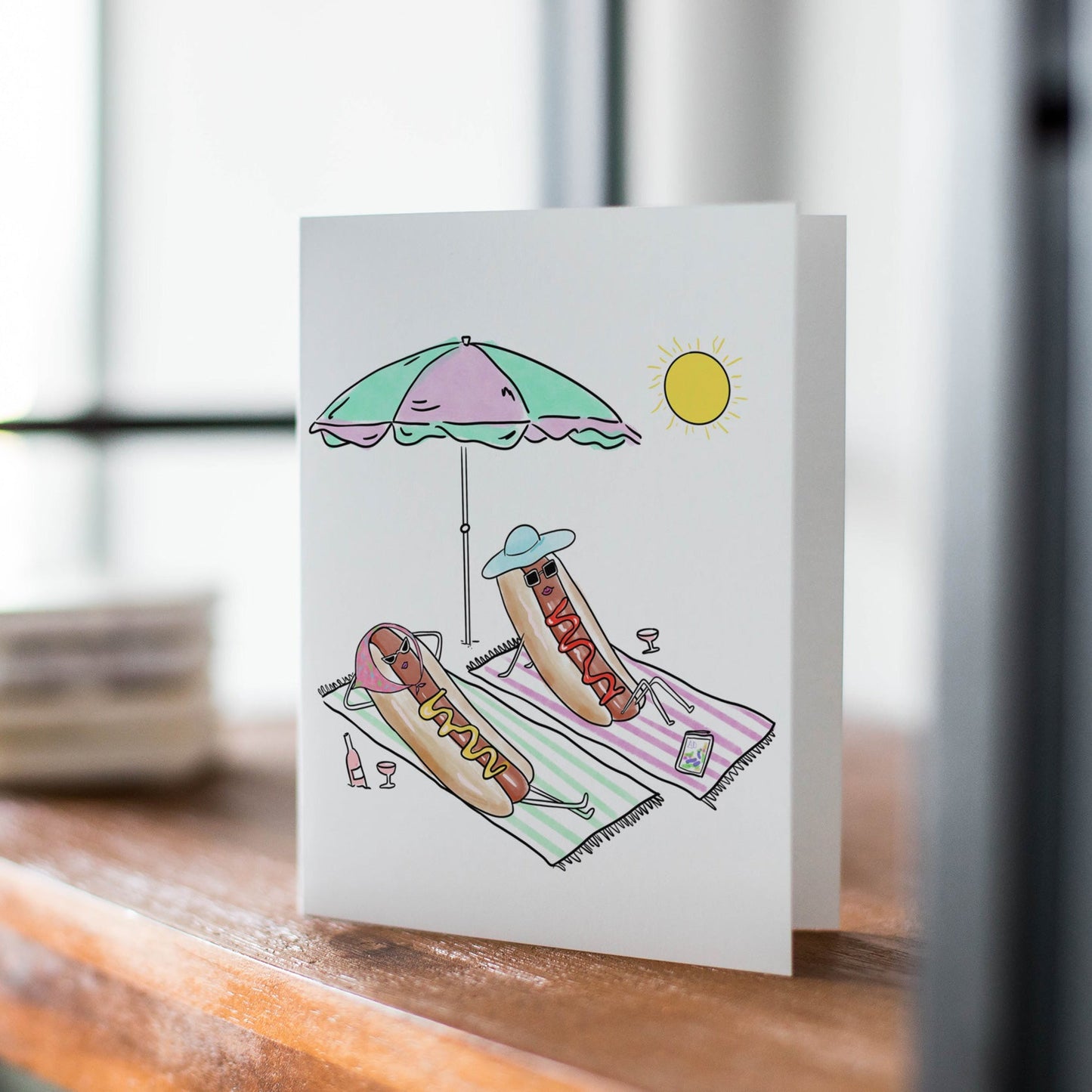 A white greeting card standing upright on a wooden table, printed with a whimsical beach scene featuring a hotdog under a pastel umbrella and lounge chair.
