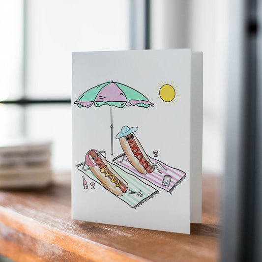 A white greeting card standing upright on a wooden table, printed with a whimsical beach scene featuring a hotdog under a pastel umbrella and lounge chair.
