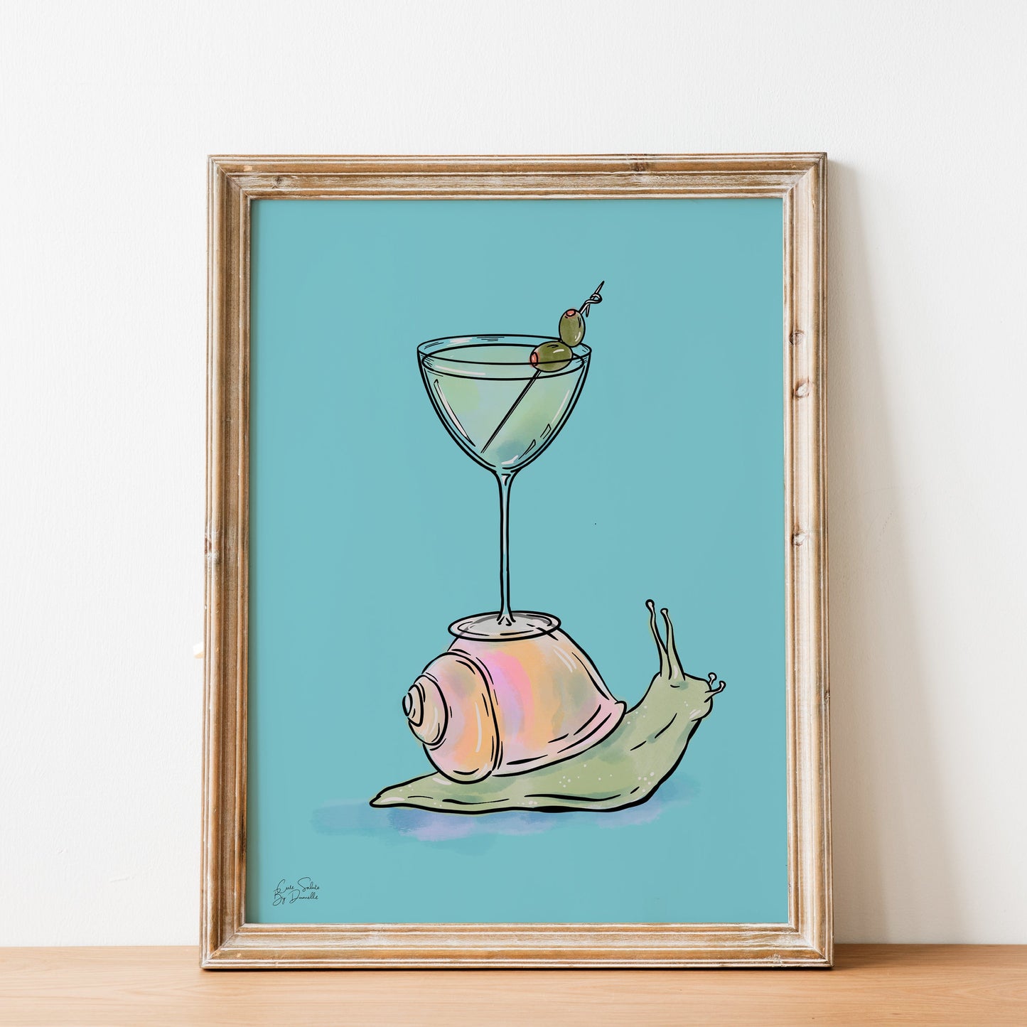 A framed art print featuring a whimsical illustration of a pink and green snail carrying a martini glass on its shell, set against a light blue background and displayed on a wooden surface against a white wall.