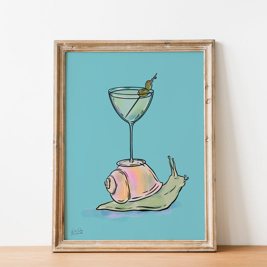 A framed art print featuring a whimsical illustration of a pink and green snail carrying a martini glass on its shell, set against a light blue background and displayed on a wooden surface against a white wall.
