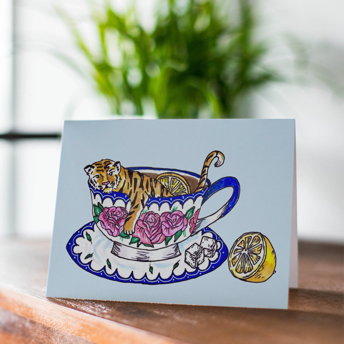 A A4 greeting card with a whimsical illustration of a tiger sitting inside a floral teacup on a saucer, displayed on a wooden surface with a blurred window and plant in the background.