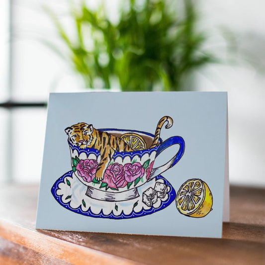 A A4 greeting card with a whimsical illustration of a tiger sitting inside a floral teacup on a saucer, displayed on a wooden surface with a blurred window and plant in the background.