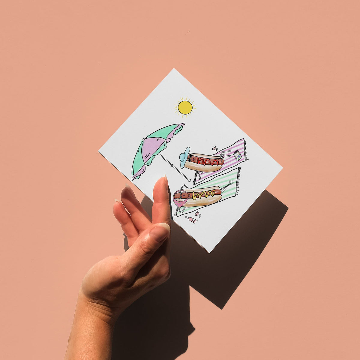 A hand holds a small square white card  featuring an illustration of two whimsical hot dogs lounging in the sun, against a solid peach-pink background. The card is angled, casting a strong shadow on the surface below.