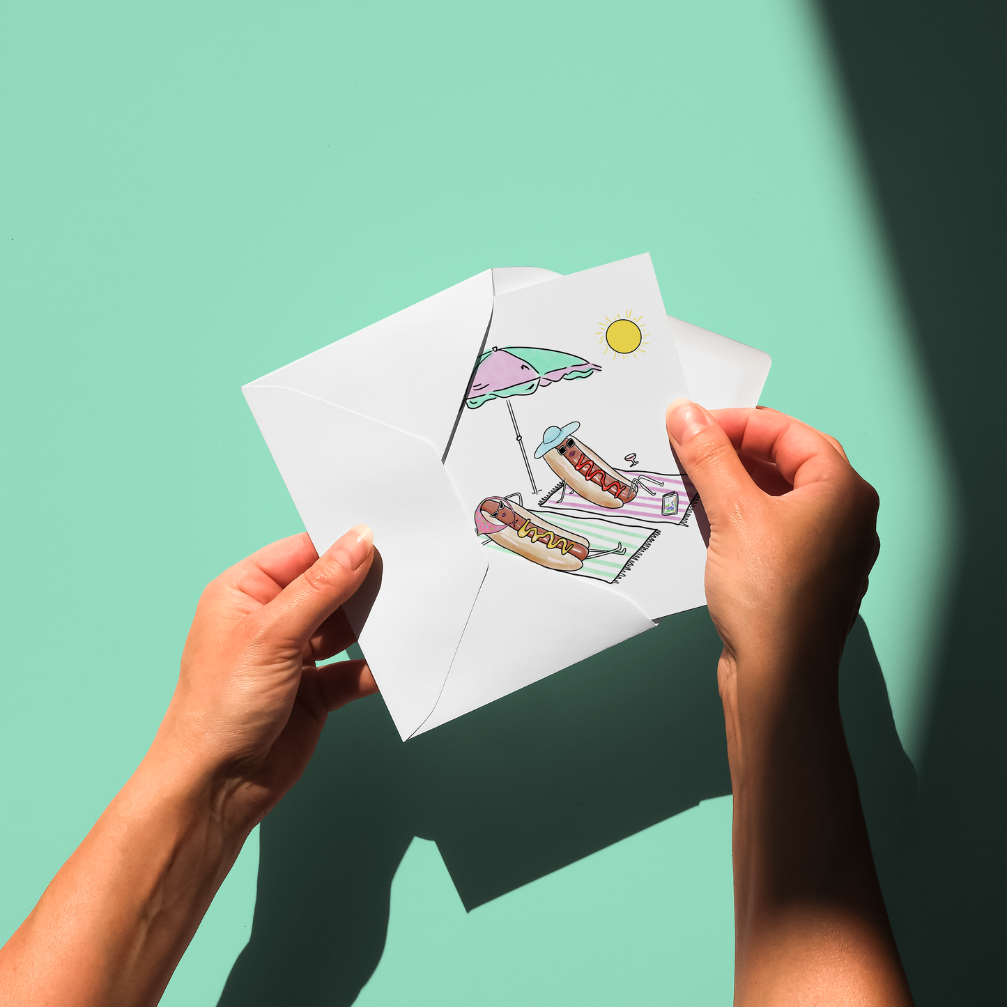 Two hands holding a folded white paper card or envelope against a mint green background. The card has a colorful illustration printed on it, showing a beach umbrella, a hot dog, a drink, and a yellow sun.