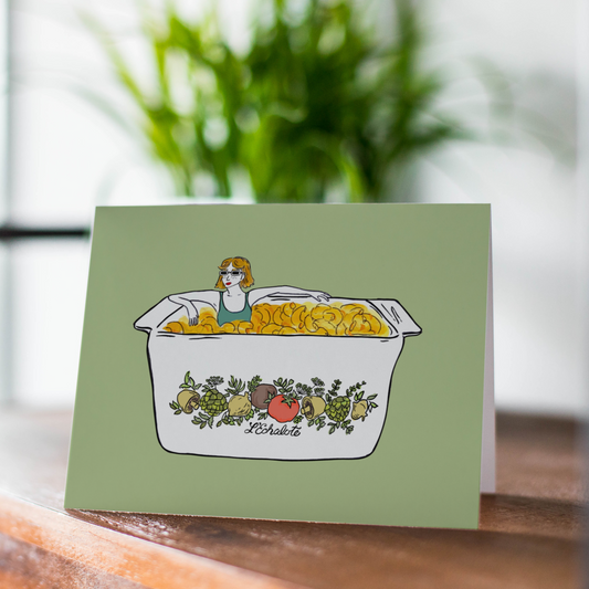 Card with illustration of a person in a bathtub filled with mac n cheese surrounded by fruits and vegetables on a wooden surface.